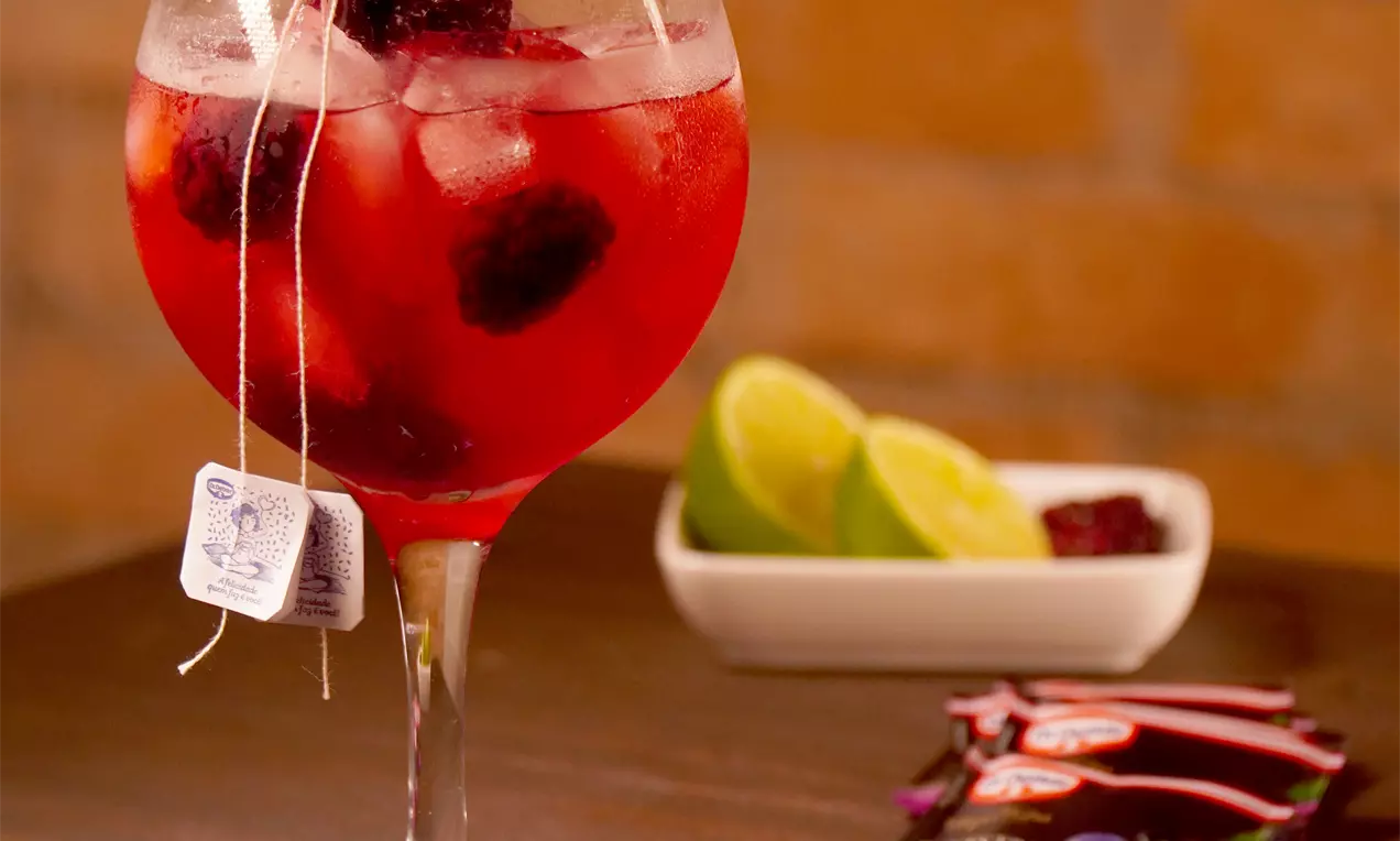 Gin tônica com chá de frutas vermelhas em copo alto com gelo, fatia de limão e bagas, tom rosado translúcido
